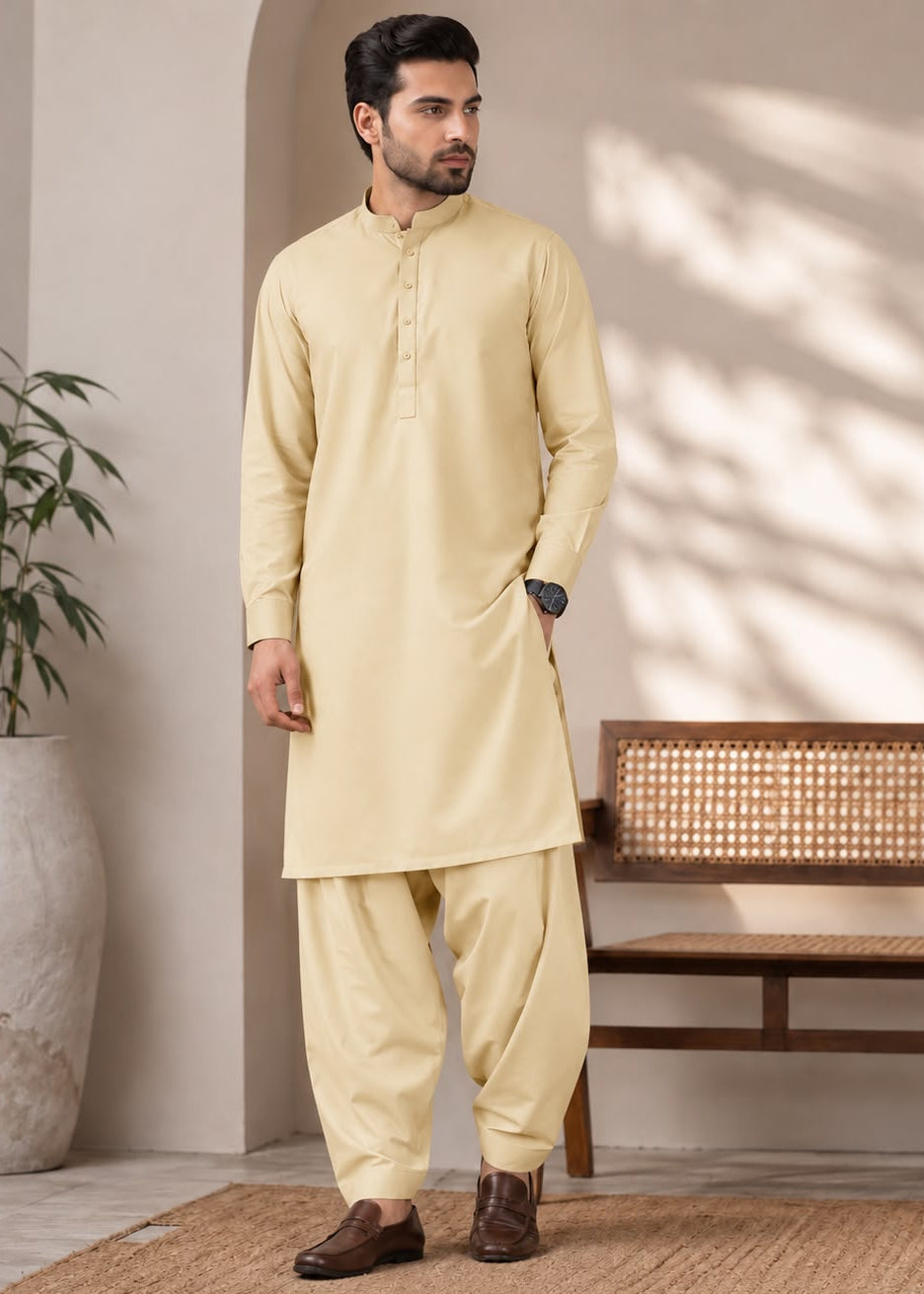 A man modeling a light beige kurta and pant set in a styled indoor setting with a bench, potted plant, and textured rug.