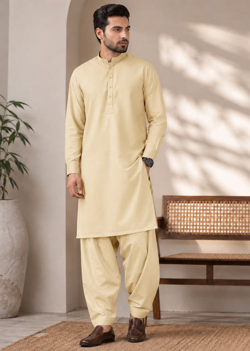 A man modeling a light beige kurta and pant set in a styled indoor setting with a bench, potted plant, and textured rug.
