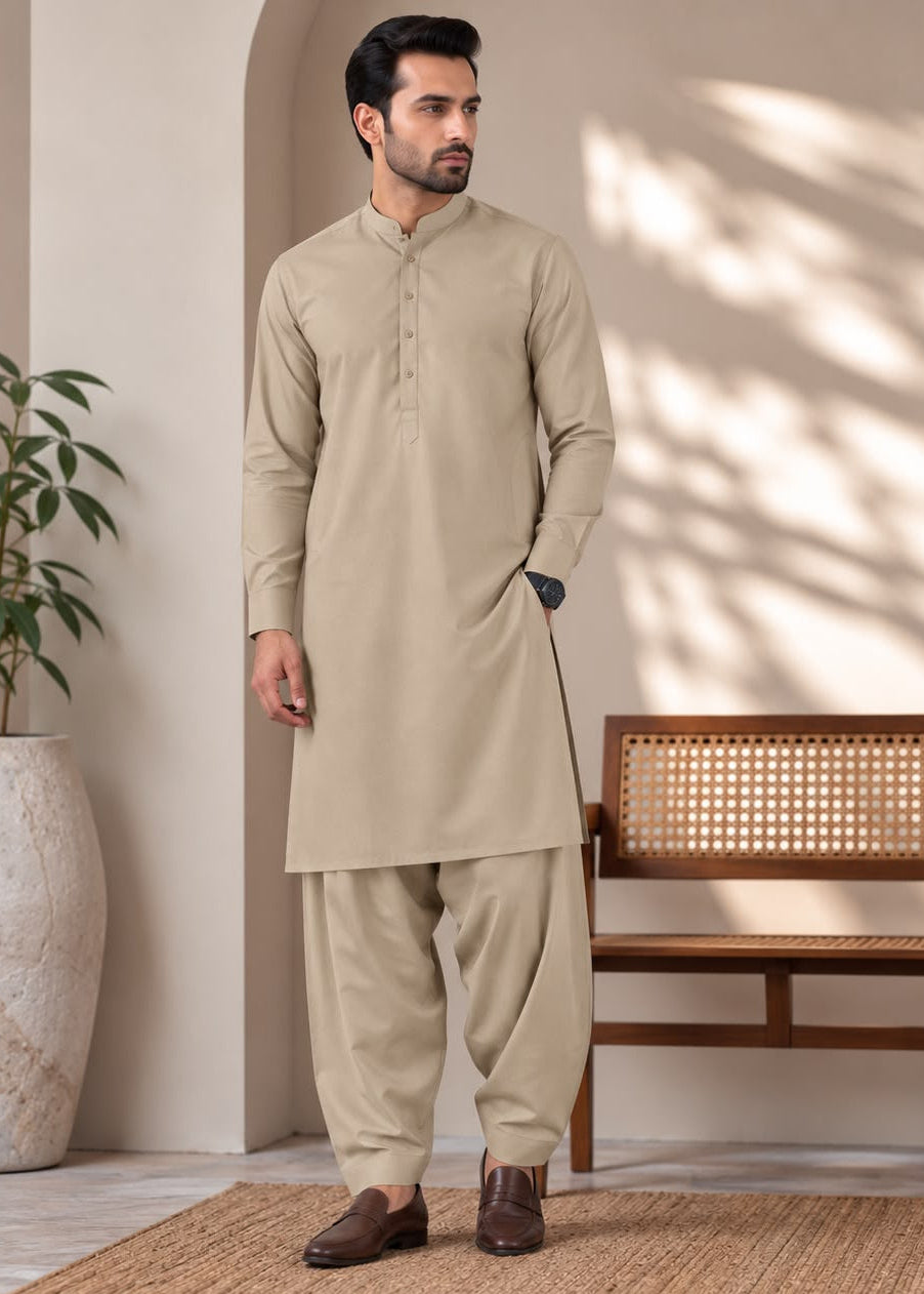 A man wearing a light beige traditional kurta and pant set stands in a styled indoor setting with a wooden bench, potted plant, and warm sunlight shadows on the wall.