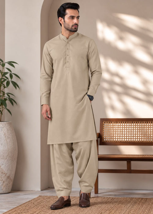 A man wearing a light beige traditional kurta and pant set stands in a styled indoor setting with a wooden bench, potted plant, and warm sunlight shadows on the wall.
