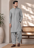 A man modeling a light gray three-piece traditional outfit consisting of a long-sleeve kurta, matching trousers, and brown leather shoes, standing in a modern, sunlit room with a bench and potted plant.