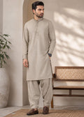 A man modeling a light beige kurta and pant set in a styled indoor setting with a bench, potted plant, and soft shadow patterns on the wall.