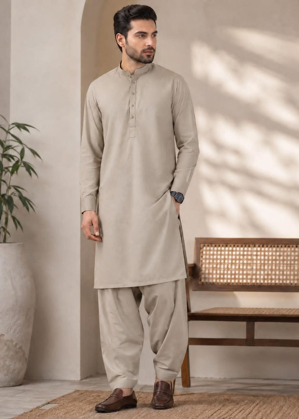 A man modeling a light beige kurta and pant set in a styled indoor setting with a bench, potted plant, and soft shadow patterns on the wall.