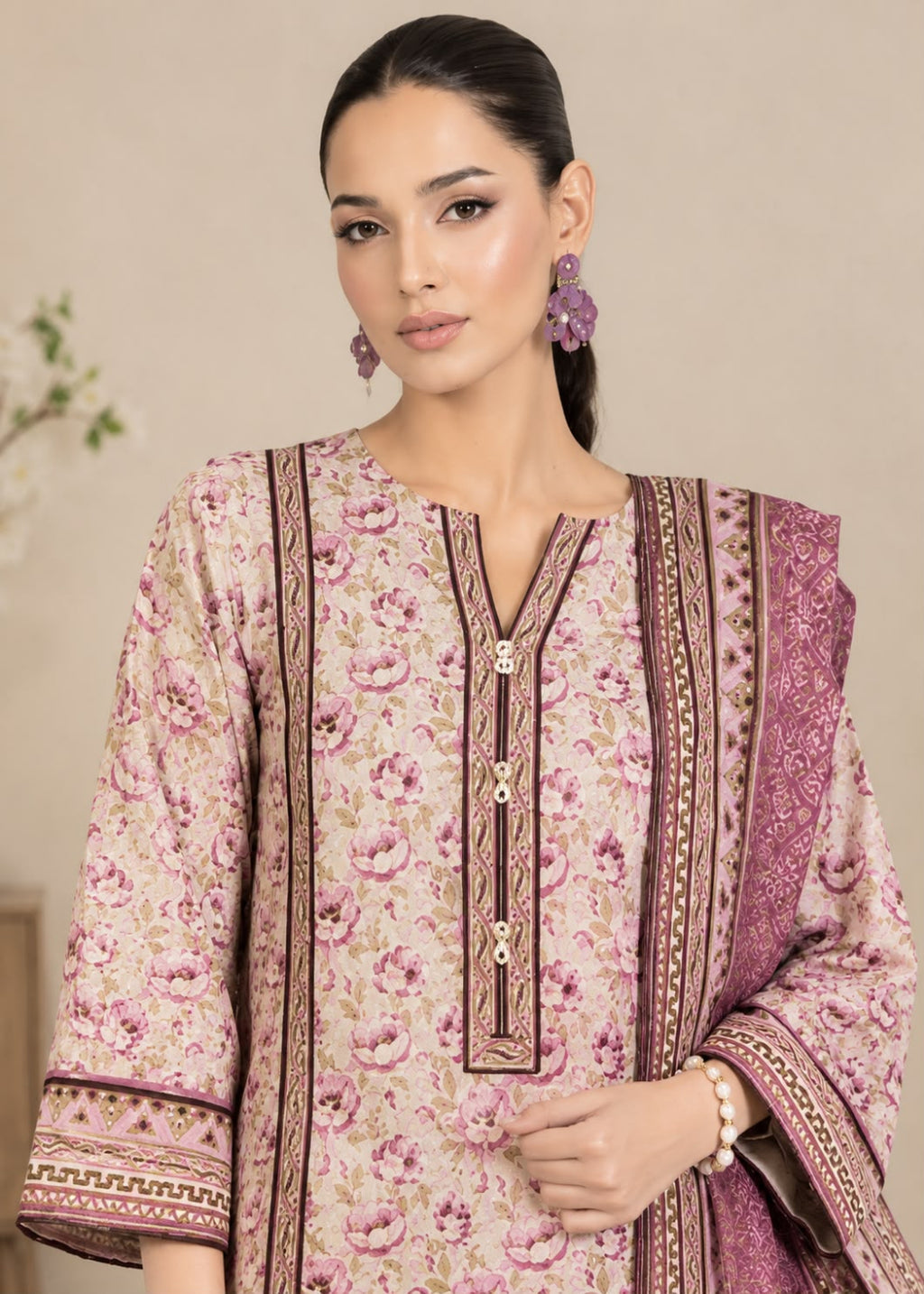 A woman modeling a traditional three-piece salwar kameez suit in a dusty pink and purple floral print. The kurta features a V-neckline with gold-tone decorative buttons, and the matching dupatta and trousers are decorated with detailed purple and gold borders.