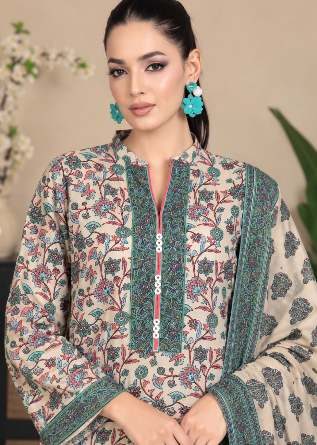 A woman wearing a beige printed kurta with teal and red floral patterns, a matching dupatta, and turquoise earrings, posed against a softly blurred indoor background.