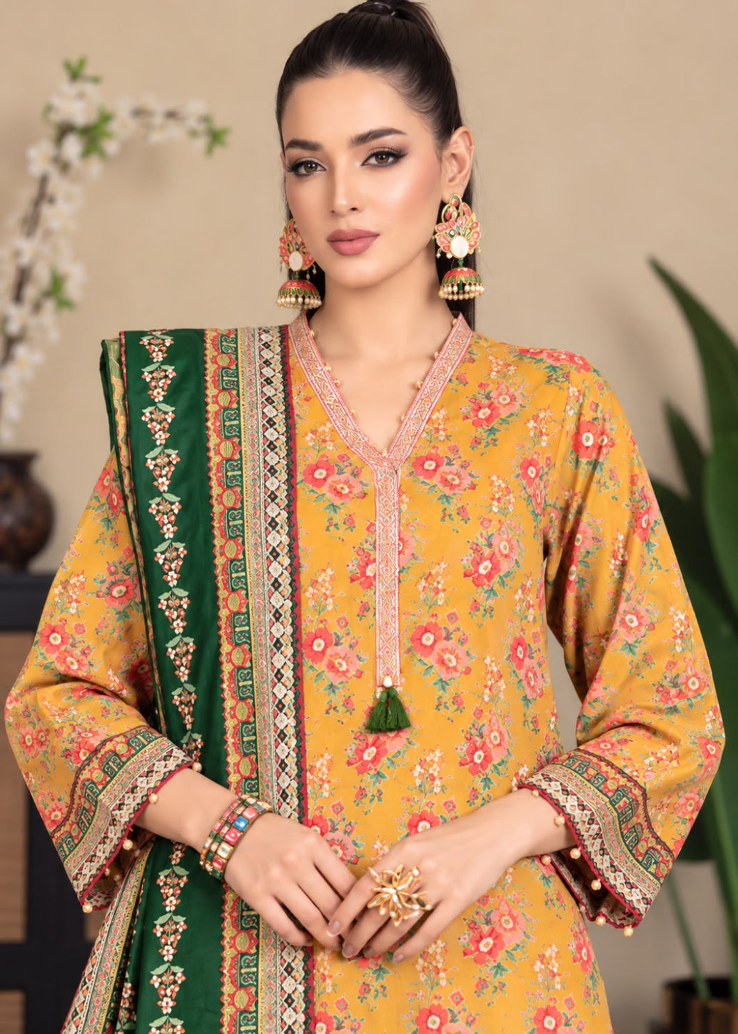 A woman models a mustard yellow floral printed salwar kameez suit with a green dupatta featuring intricate gold and red borders, styled with traditional gold jewelry against a warm beige background.