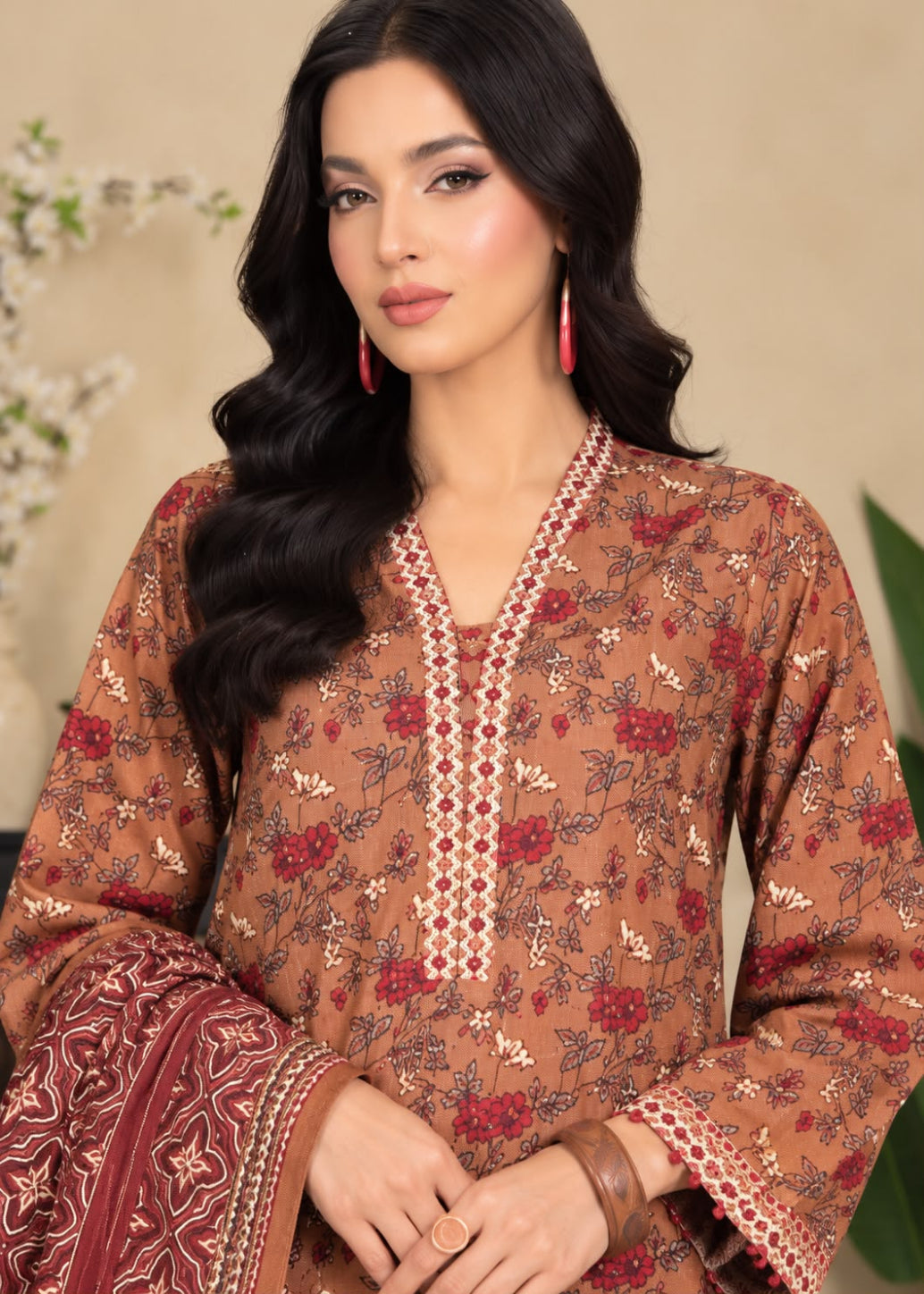 A woman modeling a brown printed salwar kameez suit with red floral motifs and a matching patterned dupatta, set against a soft beige background.