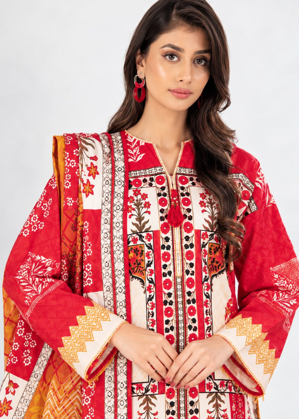 A woman modeling a red and cream printed traditional outfit with ornate embroidery, featuring a red dupatta, set against a plain light gray background.