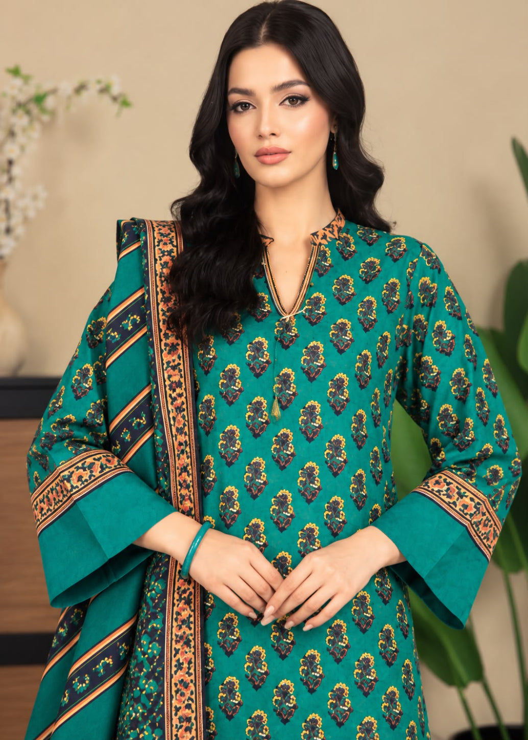 A woman modeling a teal green unstitched salwar kameez suit with a matching dupatta. The kurta features an all-over gold and black floral block print, and the sleeves and border of the dupatta have matching ornate gold and black patterns.