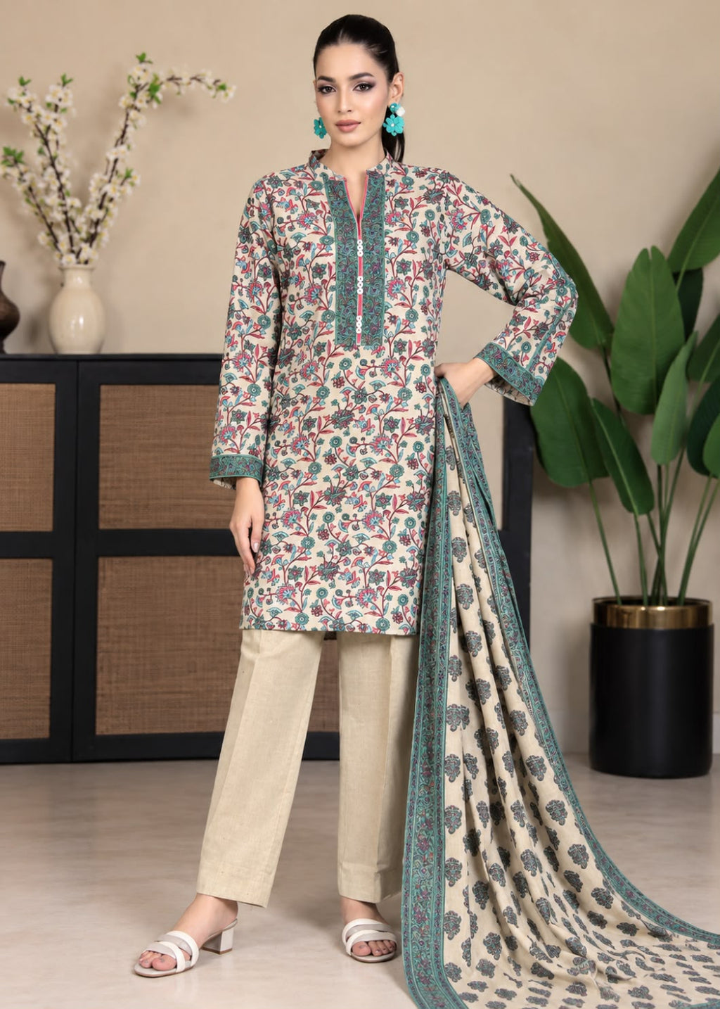 A woman models a three-piece salwar kameez suit set featuring a beige kurta with a multicolor floral print and teal borders, paired with beige trousers and a matching printed dupatta, in a styled indoor setting.