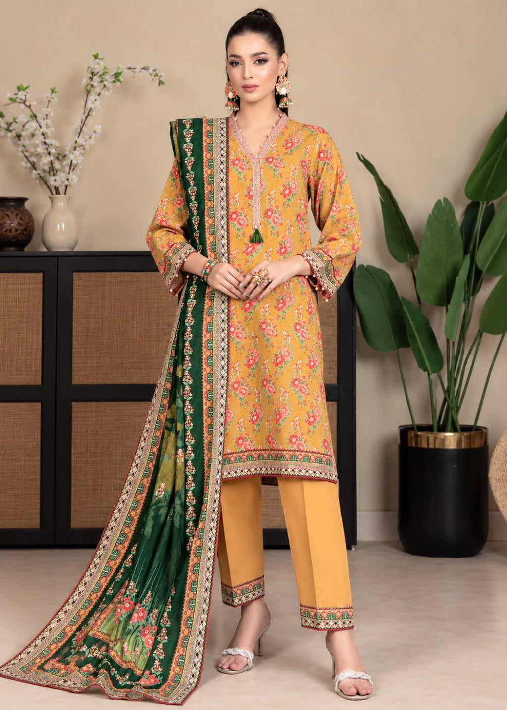 A woman models a traditional three-piece salwar kameez suit set. The set features a mustard yellow printed kurta with pink and green floral motifs, matching yellow trousers, and a dark green printed dupatta with a detailed gold and pink border.