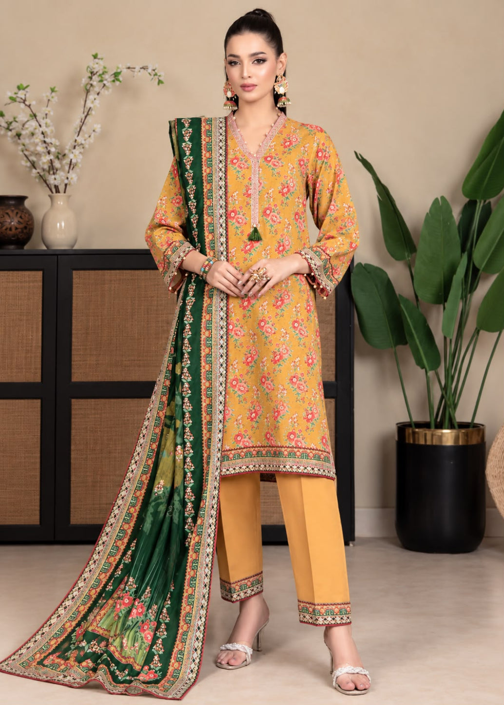 A woman models a traditional three-piece salwar kameez suit set. The set features a mustard yellow printed kurta with pink and green floral motifs, matching yellow trousers, and a dark green printed dupatta with a detailed gold and pink border.