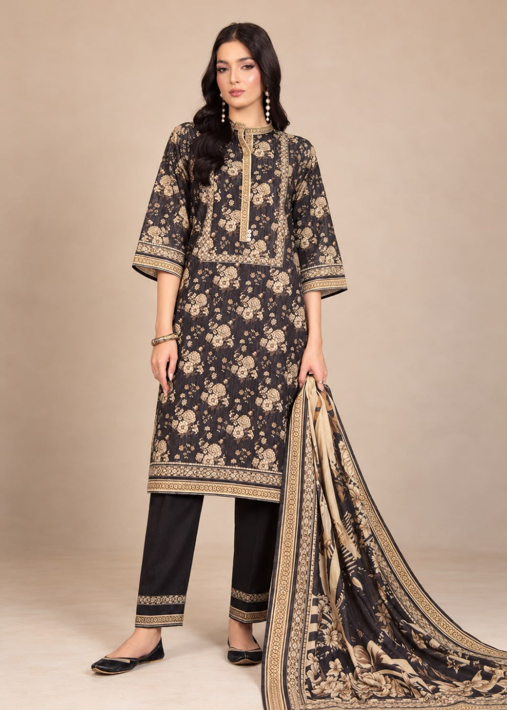 A woman models a traditional three-piece salwar kameez suit in black with beige floral prints and borders, paired with black trousers and a matching printed dupatta, set against a solid beige studio background.