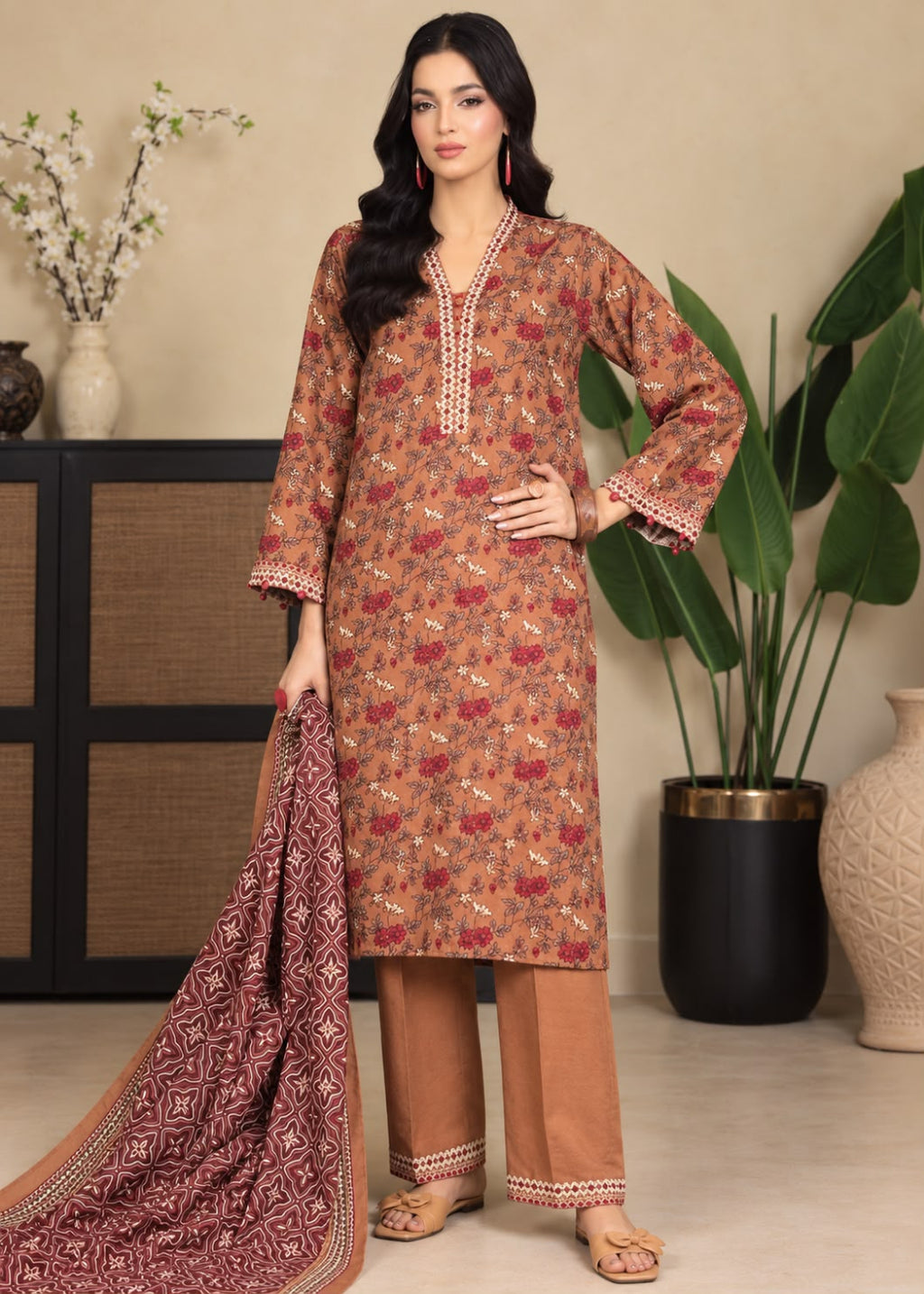 A woman models a three-piece Indian ethnic salwar kameez suit set. The set includes a rust-orange printed kurta with white embroidery on the neckline and cuffs, matching brown trousers with embroidered hems, and a dark red patterned dupatta.