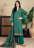A woman models a traditional South Asian salwar kameez suit in teal green with intricate gold and brown floral patterns, paired with matching wide-leg trousers and a coordinating dupatta, set against a warm beige background with decorative plants and vases.