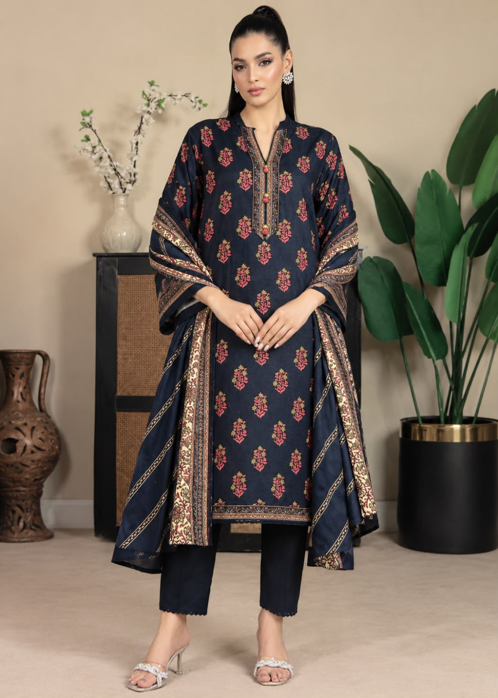 A woman models a navy blue three-piece salwar kameez suit with red and gold floral prints, paired with matching navy pants and a dupatta, in a styled indoor setting.
