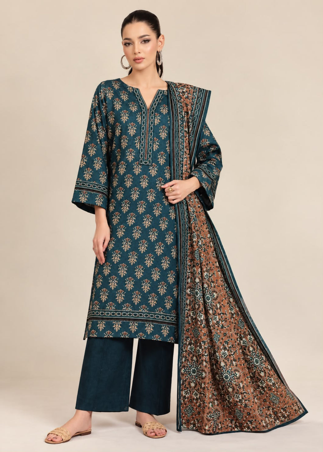 A woman modeling a traditional three-piece salwar kameez suit in teal blue, featuring a printed kurta, matching trousers, and a dupatta with brown and gold floral patterns, set against a plain beige studio background.