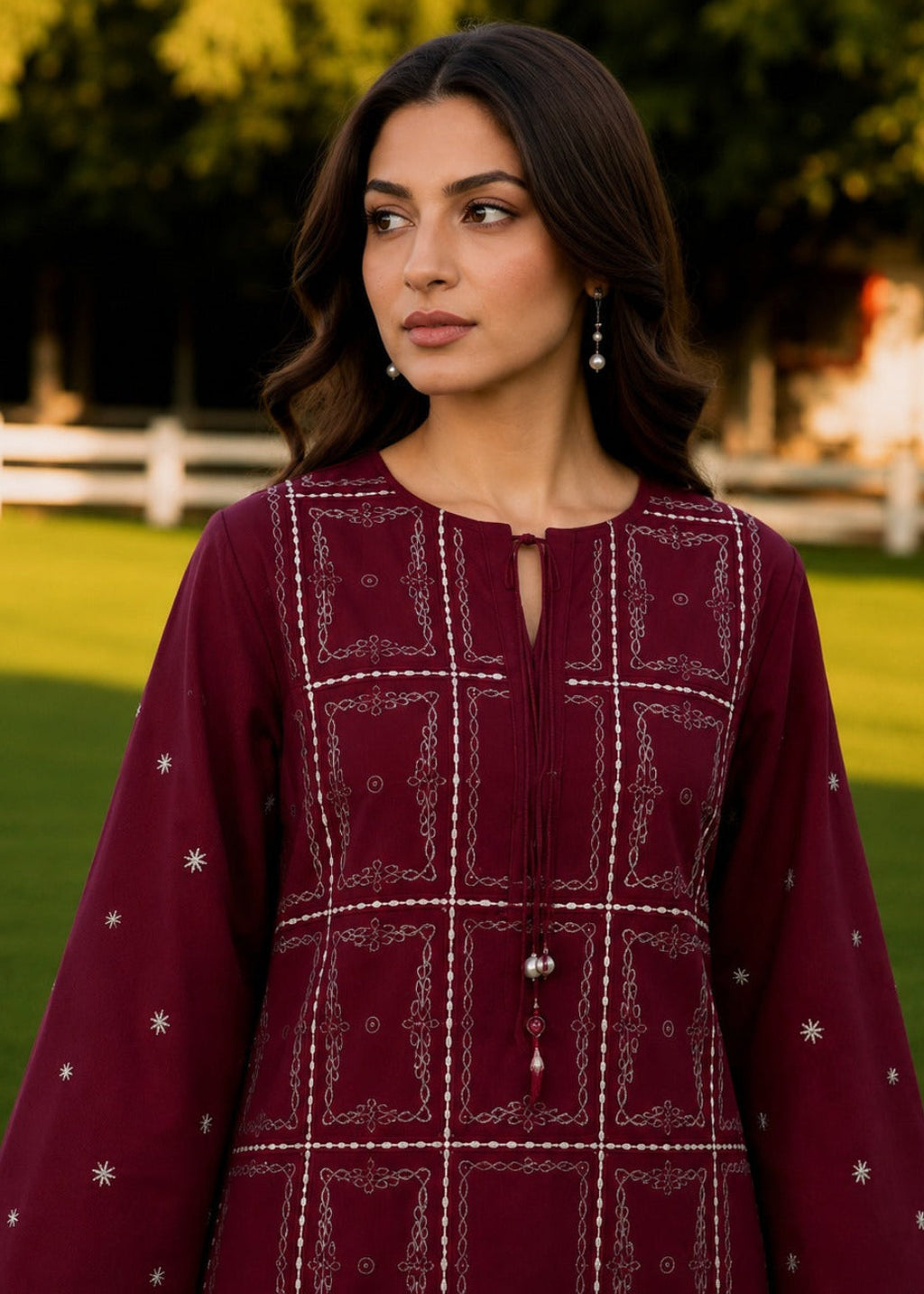 A woman wearing a deep maroon long-sleeve kurta with white geometric embroidery and star motifs, standing in a grassy park with trees in the background.