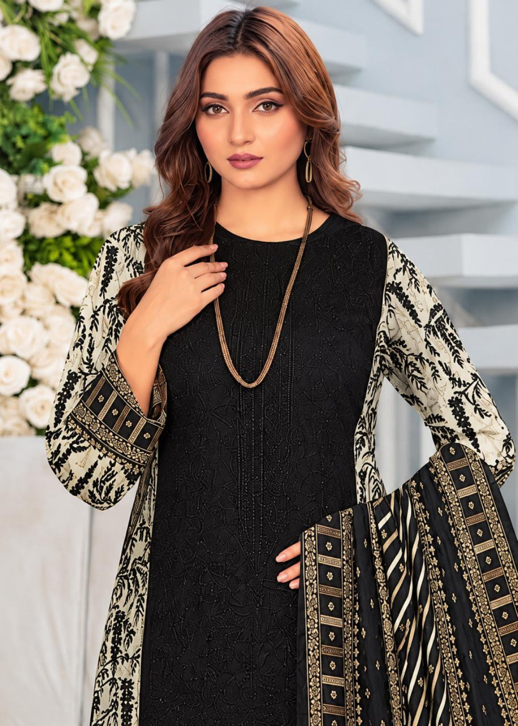 A woman models a black embroidered kurta with beige and black patterned sleeves and a matching dupatta, styled with a long gold-toned necklace against a light gray background with white flowers.