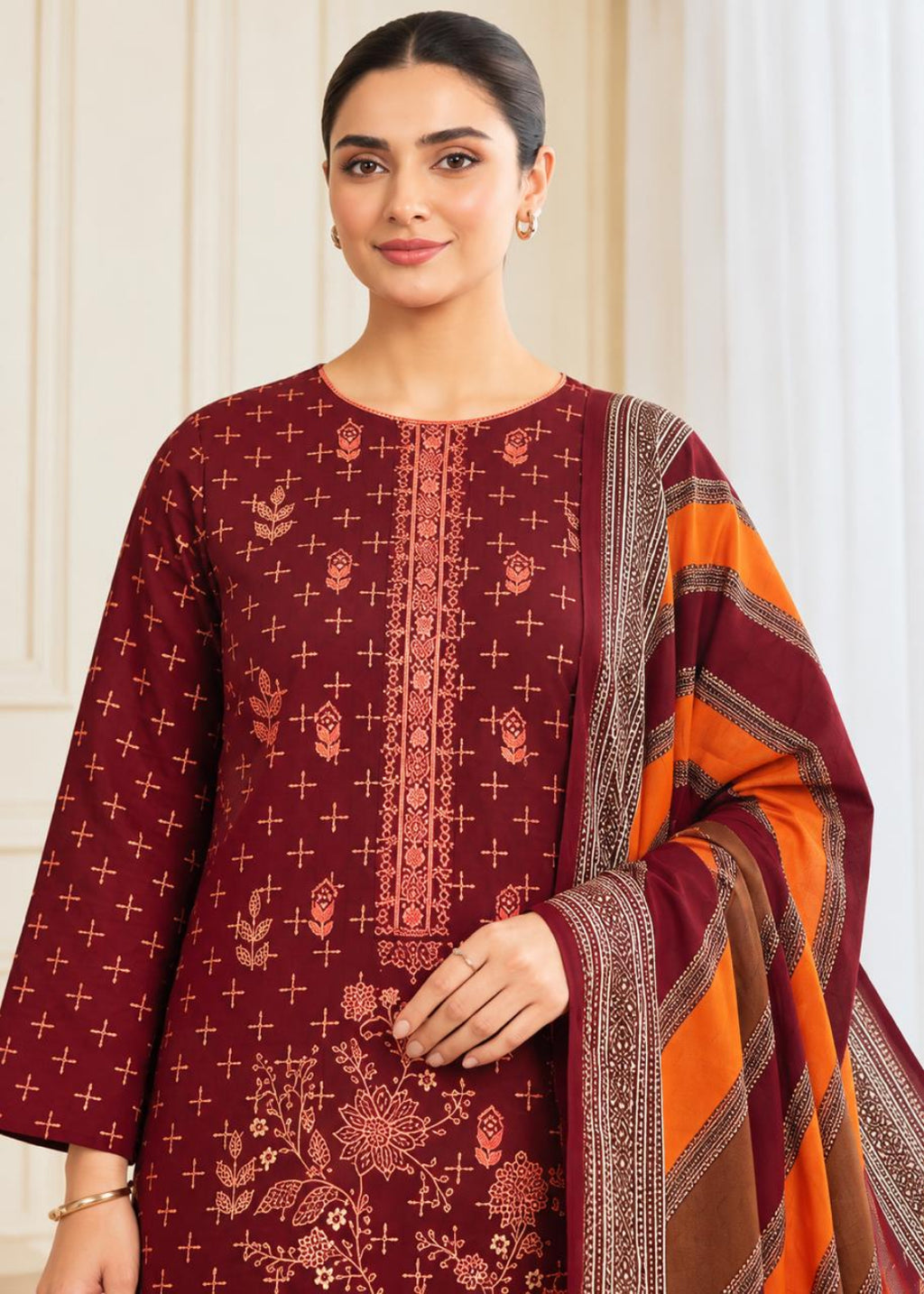 A woman modeling a deep maroon three-piece salwar kameez suit with matching printed fabric and a contrasting orange and maroon dupatta, set against a soft beige indoor background.