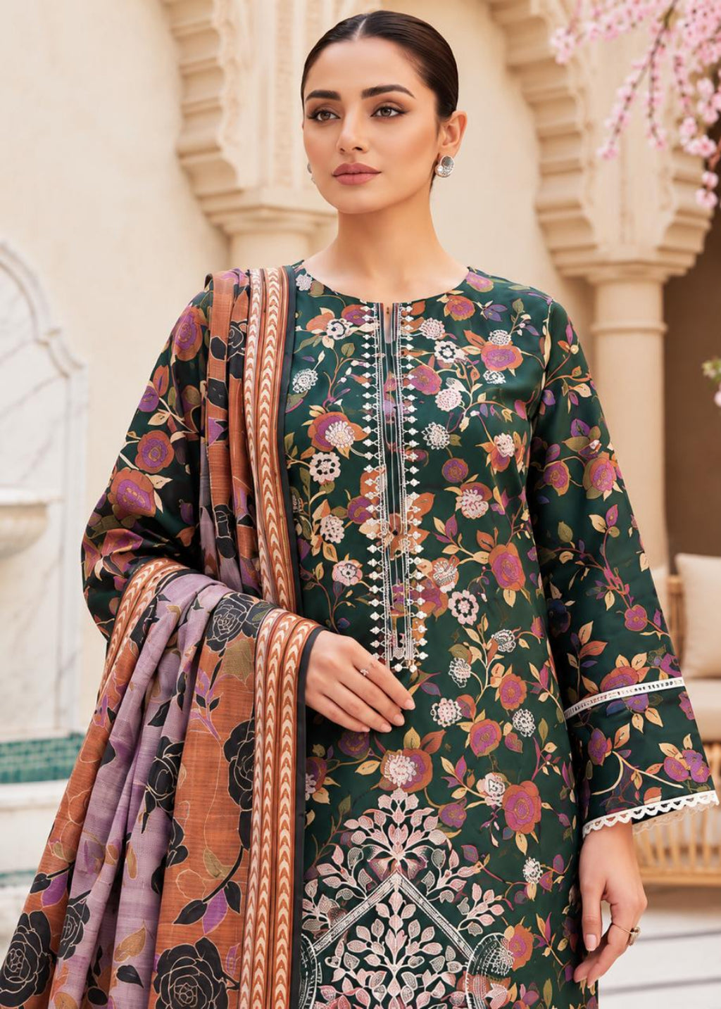 A woman models a dark green floral printed salwar kameez suit with a matching dupatta in a courtyard setting. The kurta features a floral pattern in pink, orange, and white, with silver mirror work embroidery down the center front.