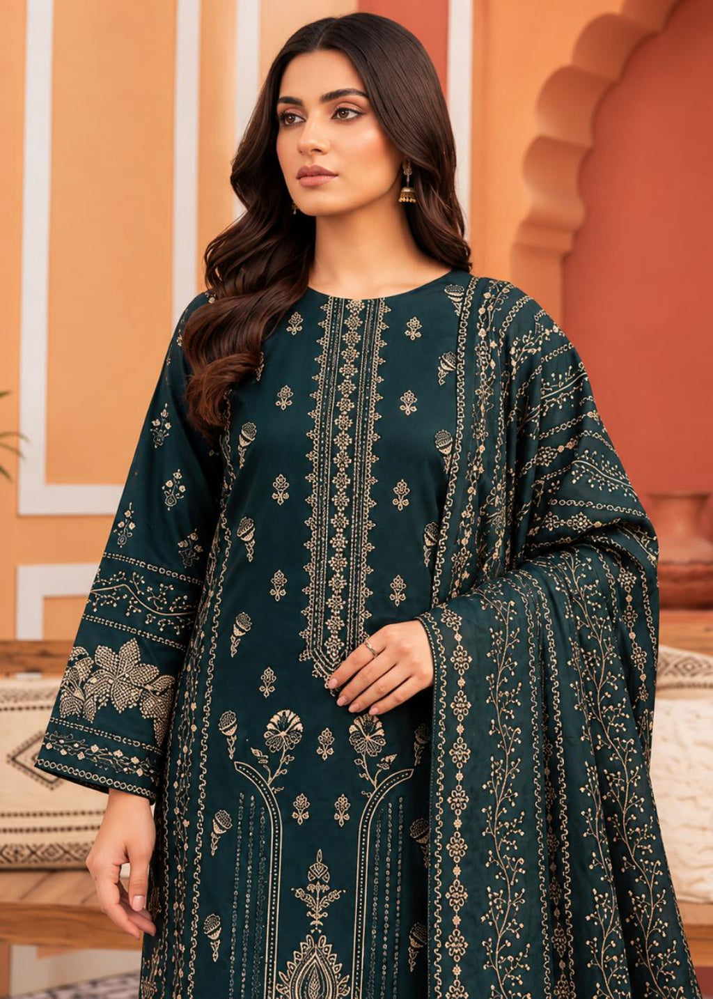 A woman modeling a dark teal green traditional South Asian salwar kameez suit with intricate gold and beige embroidery, paired with a matching dupatta, set against a warm architectural background.