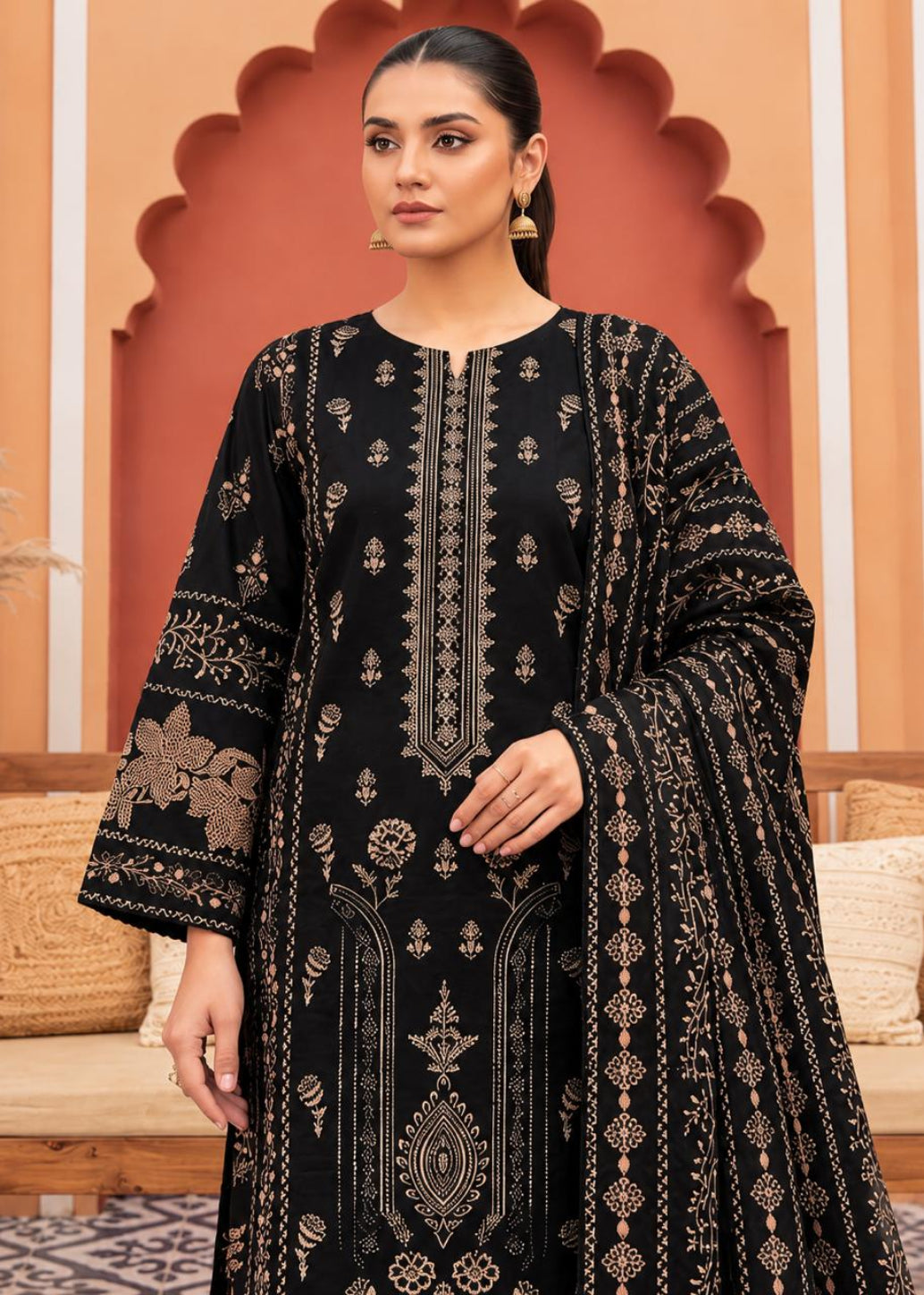 A woman modeling a black traditional outfit with intricate beige and gold embroidery, set against a warm architectural background with arches and cushions.