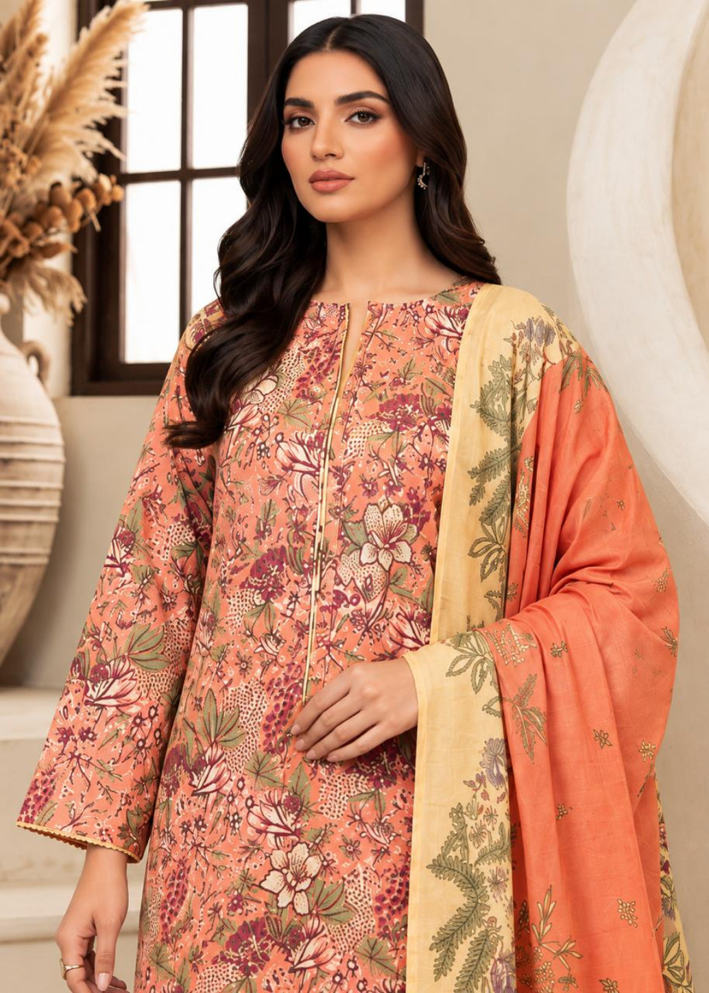 A woman modeling a peach and orange floral printed salwar kameez suit with a beige dupatta featuring green leafy patterns, set against a warm beige interior background.
