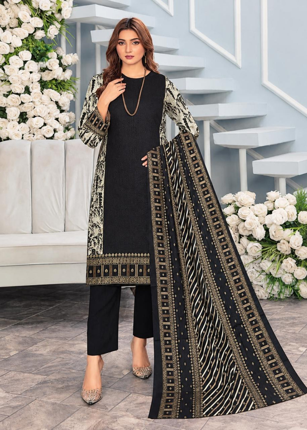 A woman models a three-piece black salwar kameez suit set with beige and gold geometric patterns. The set includes a long tunic with a printed kameez collar, matching printed sleeves, straight-fit pants, and a long dupatta draped over her shoulder.