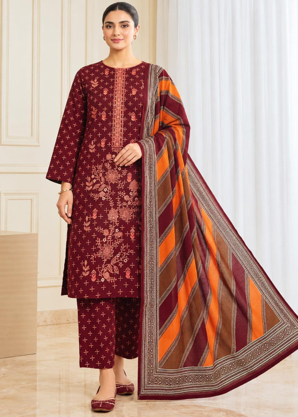 A woman models a traditional three-piece salwar kameez suit in deep maroon with floral embroidery, paired with matching trousers and a striped orange, brown, and maroon dupatta.