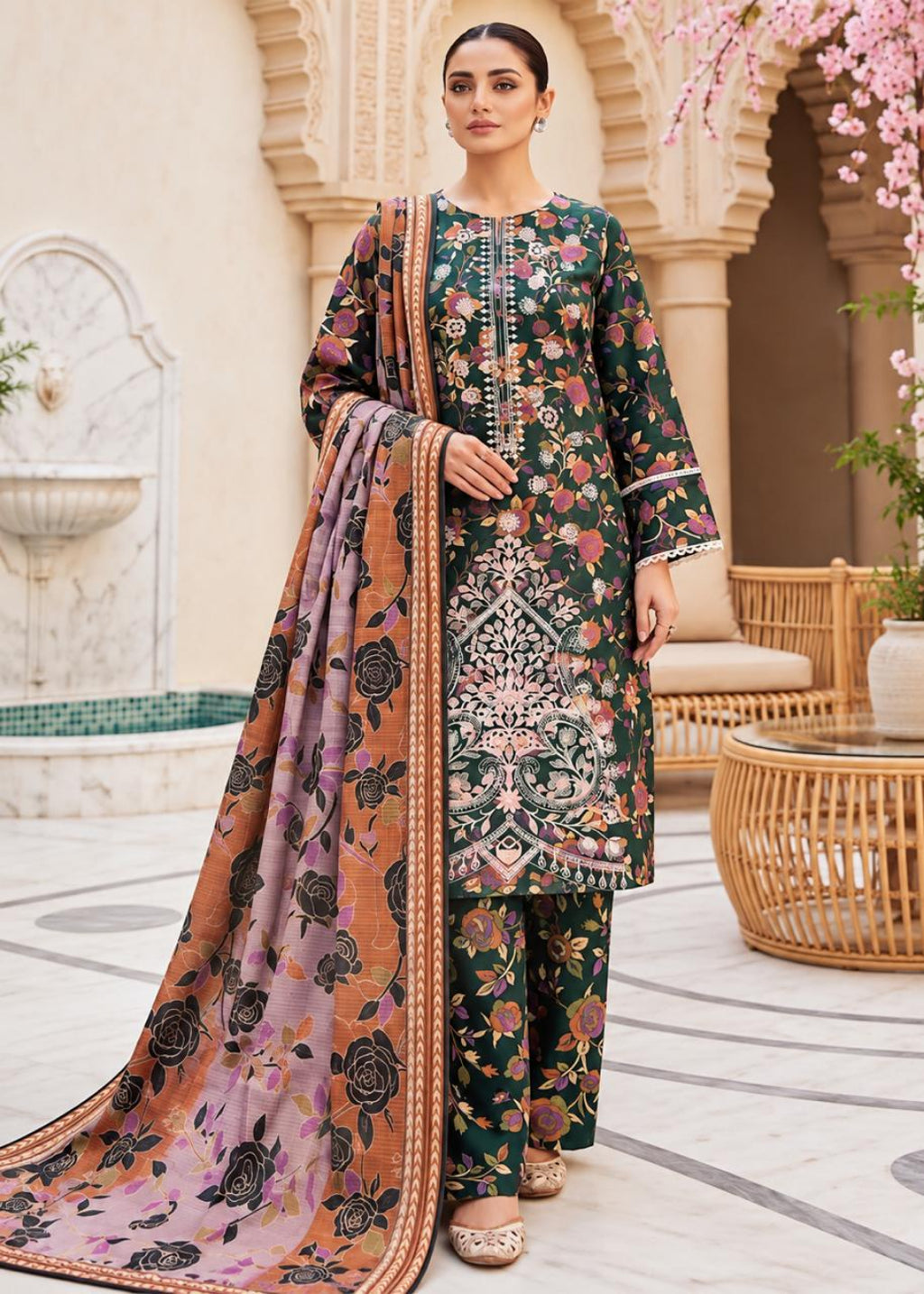 A woman models a traditional three-piece salwar kameez suit in a courtyard setting. The set features a dark green base with intricate floral prints in pink, orange, and black, paired with a matching printed dupatta in dusty pink and orange tones.