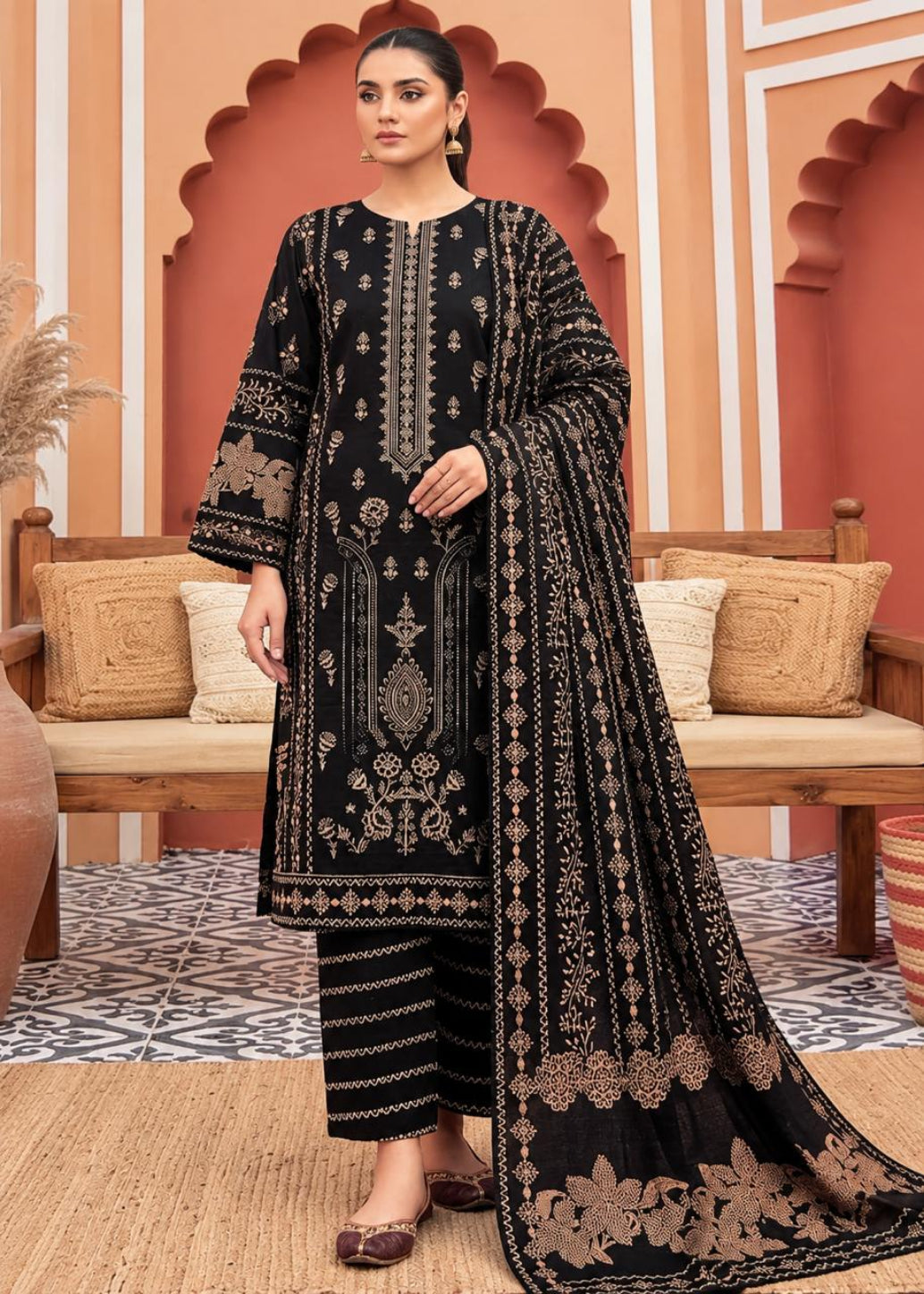 A woman models a black three-piece ethnic suit with intricate gold embroidery, featuring a long kurta, matching trousers, and a draped dupatta, set against a warm, arched architectural background.