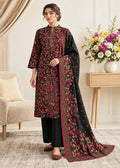 A woman models a traditional three-piece salwar kameez suit in black, featuring intricate red and gold floral embroidery, paired with black trousers and a matching embroidered dupatta, set in a warm, elegant living room.