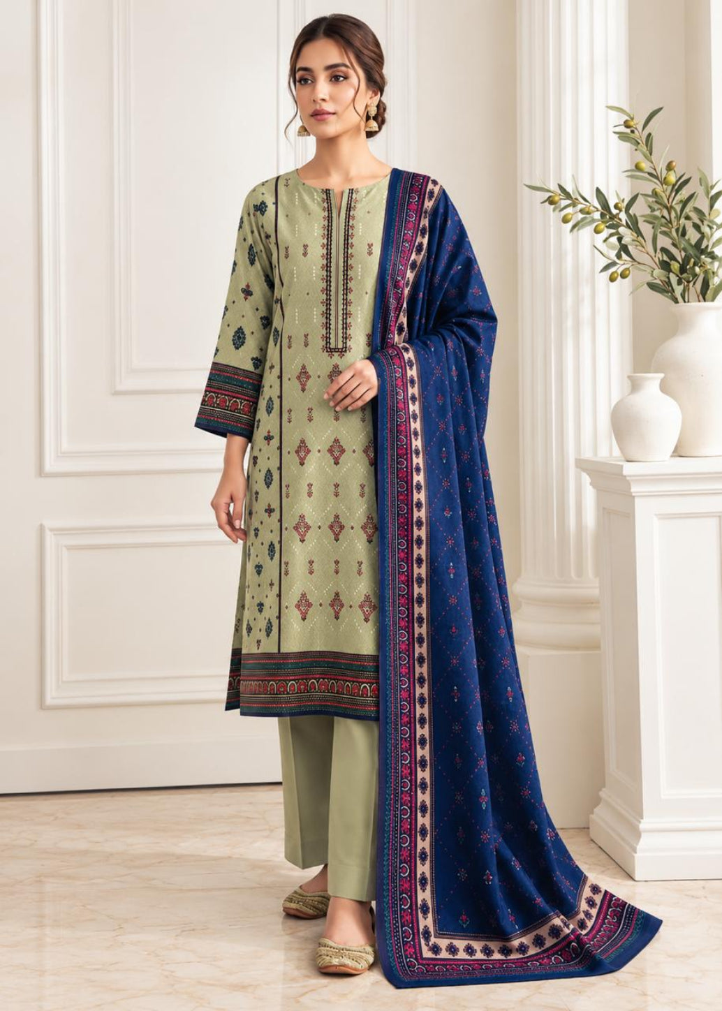 A woman models a traditional three-piece salwar kameez suit set. The kurta is sage green with intricate geometric and floral patterns in red, blue, and gold, paired with matching sage green trousers and a deep navy blue dupatta featuring a detailed multicolor border.