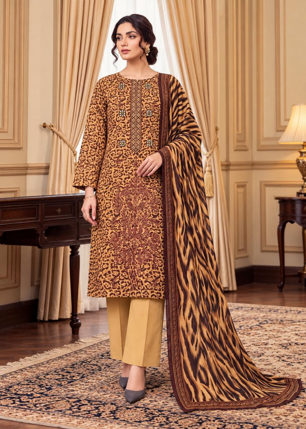 A woman models a traditional Indian salwar kameez suit set in an elegant room. The set features a brown and gold patterned kameez kurta with matching trousers and a dupatta featuring a bold tiger-stripe pattern.