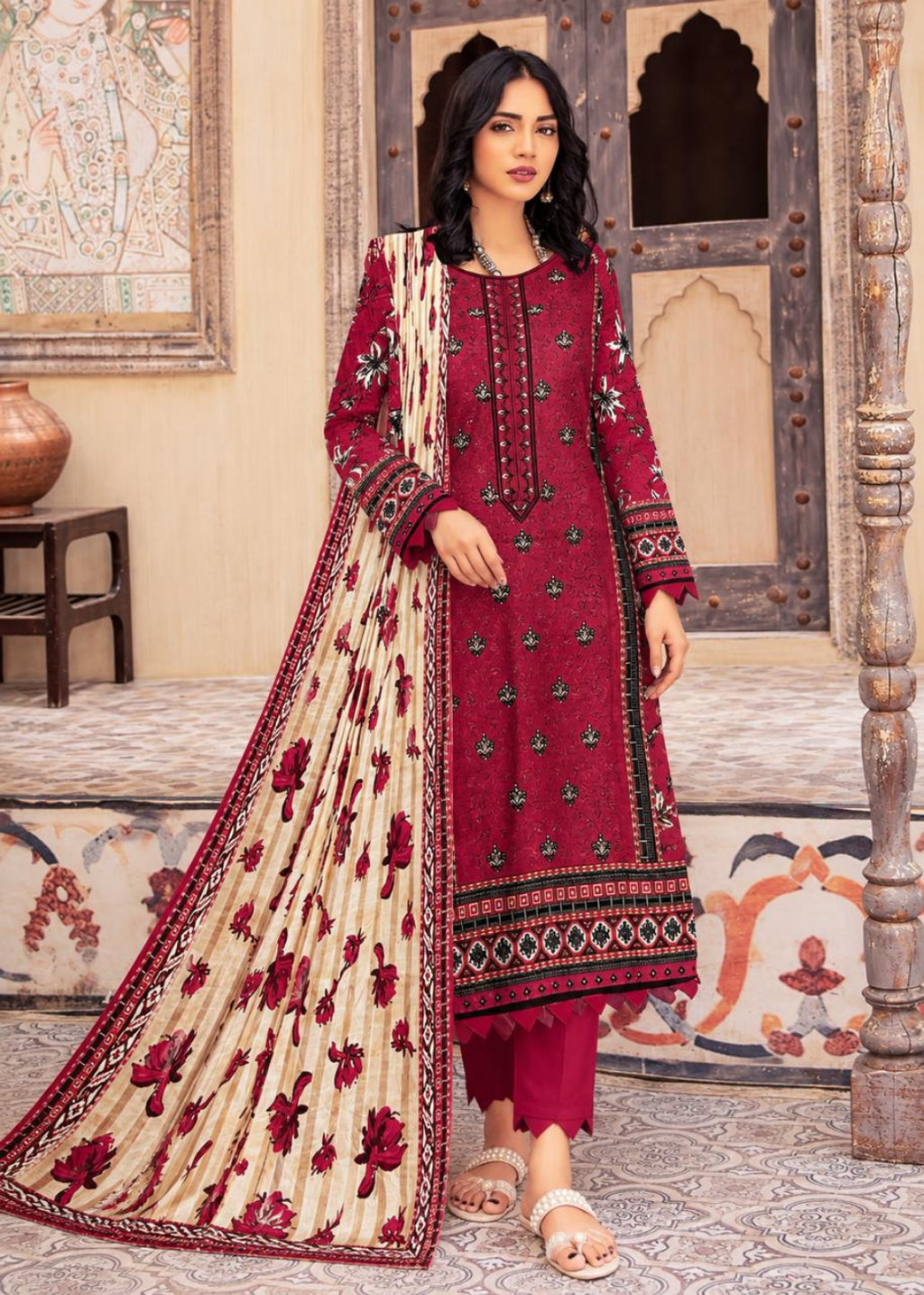 A woman models a traditional Indian salwar kameez suit in a courtyard setting. The outfit features a deep maroon red tunic with floral embroidery, matching maroon pants, and a beige dupatta with red floral motifs and a red border.