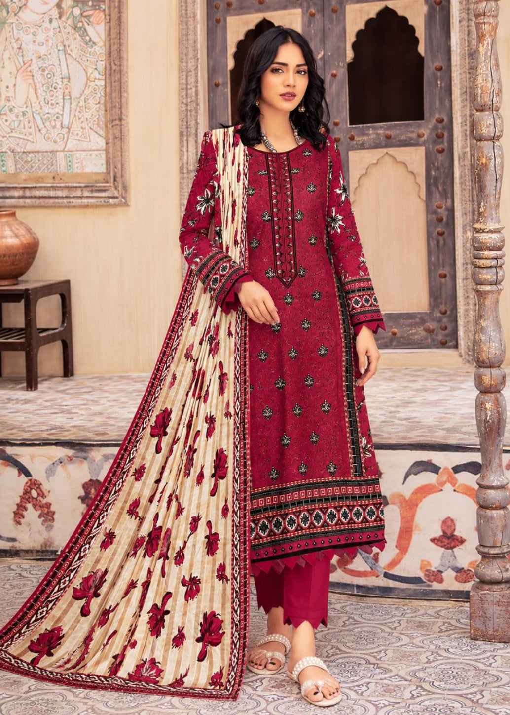 A woman models a traditional Indian salwar kameez suit in a courtyard setting. The outfit features a deep maroon red tunic with floral embroidery, matching maroon pants, and a beige dupatta with red floral motifs and a red border.