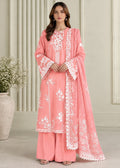 A woman modeling a pink traditional three-piece outfit with white floral embroidery and a matching dupatta, standing in front of a neutral indoor backdrop with a vase of flowers.