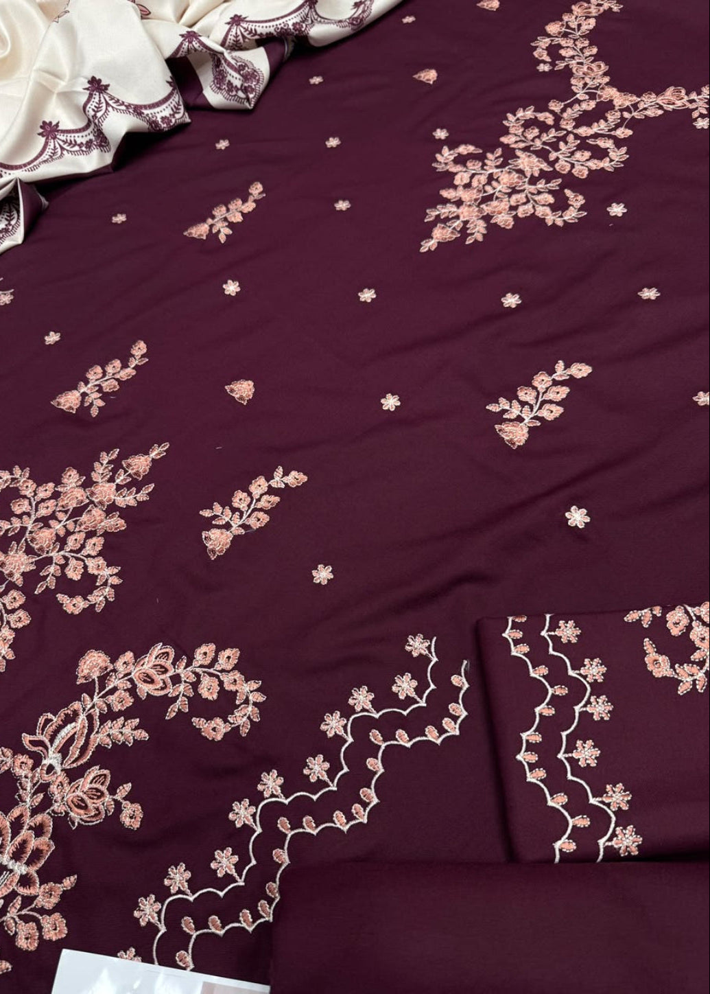 A close-up of a deep maroon fabric with intricate light pink floral embroidery and small embroidered motifs, showing folded matching fabric and a draped cream-colored embroidered textile.