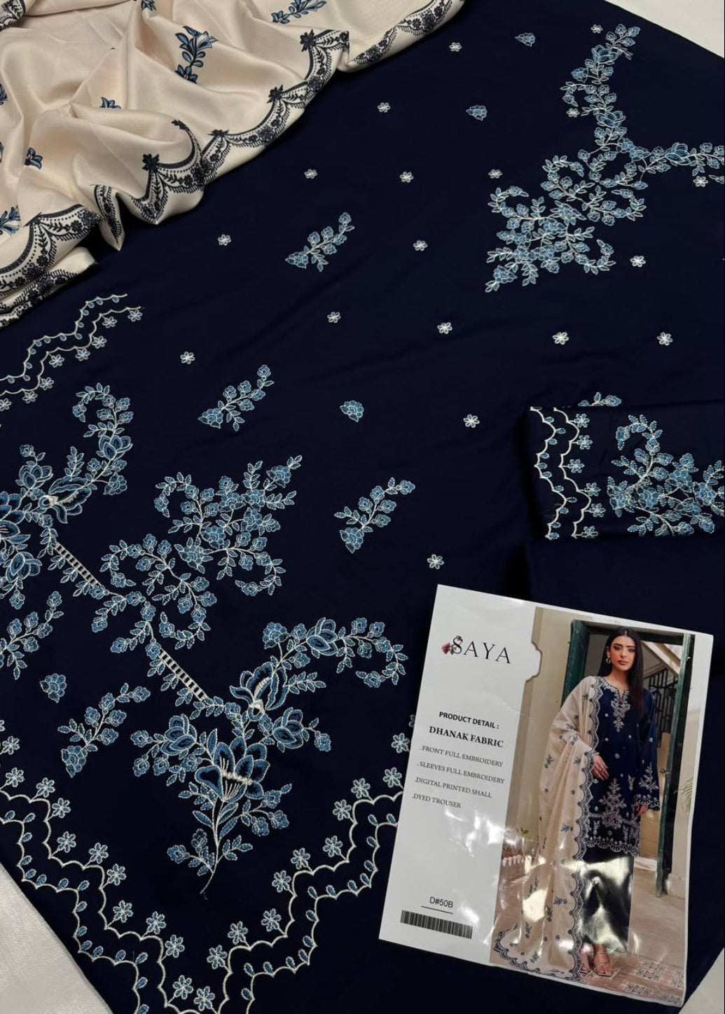 A navy blue embroidered fabric set with light blue floral embroidery, a cream dupatta, and a printed brand card showing a model. The brand name SAYA is visible on the card.