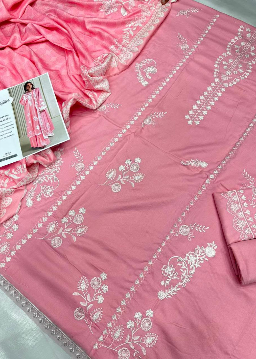 A pink traditional embroidered fabric set with white floral embroidery, shown laid out with a matching dupatta and folded fabric piece. A small printed card is visible near the upper left.