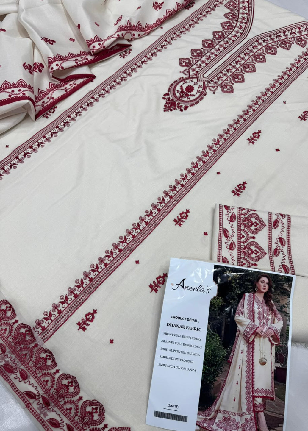 An off-white/cream embroidered fabric set with deep red border and floral embroidery, shown with a printed product card labeled 'Aneda's'.