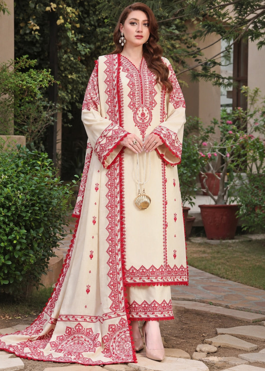 A woman stands outdoors wearing a cream-colored traditional outfit with extensive red floral embroidery. The outfit includes a long kurta with wide sleeves, matching pants, and a draped dupatta.