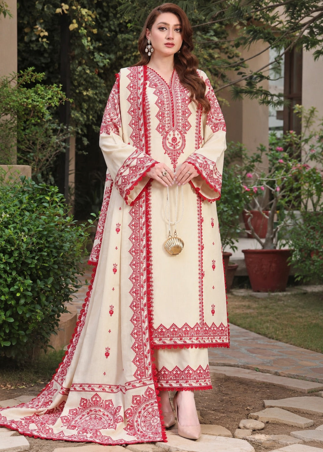 A woman stands outdoors wearing a cream-colored traditional outfit with extensive red floral embroidery. The outfit includes a long kurta with wide sleeves, matching pants, and a draped dupatta.