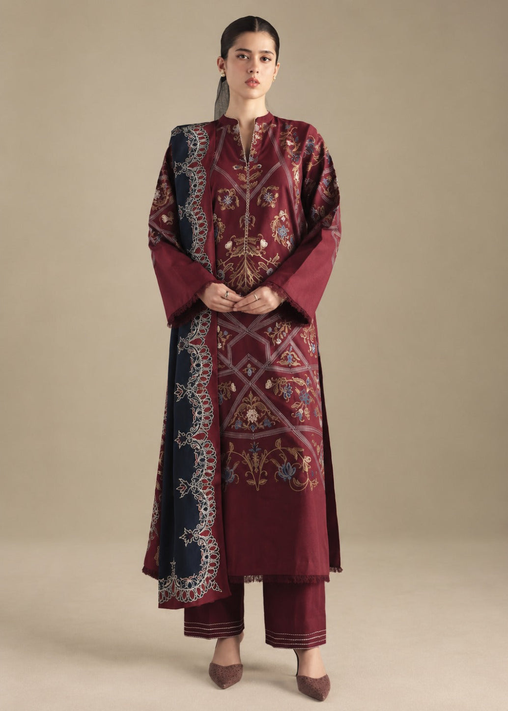 A woman modeling a traditional embroidered kurta set in deep maroon, featuring intricate gold, blue, and silver embroidery, paired with matching maroon pants, against a solid beige studio background.