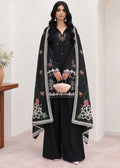 A woman models a black traditional outfit with wide-leg pants, a matching embroidered inner dress, and a large draped black shawl featuring white border trim and floral embroidery in pink, green, and white. She holds a small pink handbag in a neutral indoor setting with beige walls and tiled flooring.