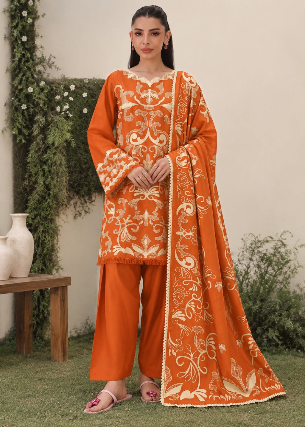 A woman models an orange traditional outfit with intricate beige embroidery, matching orange pants, and a coordinating embroidered dupatta, set against an outdoor garden backdrop.