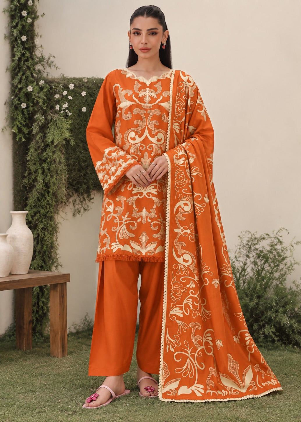 A woman models an orange traditional outfit with intricate beige embroidery, matching orange pants, and a coordinating embroidered dupatta, set against an outdoor garden backdrop.