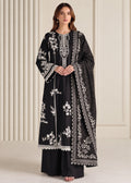 A woman models a black traditional outfit with intricate white floral embroidery and silver mirror work, paired with a matching embroidered dupatta, set against a cream-colored paneled background.