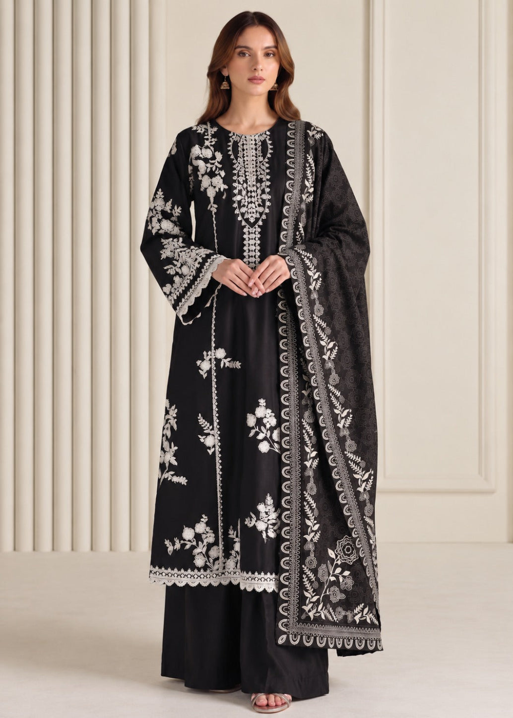 A woman models a black traditional outfit with intricate white floral embroidery and silver mirror work, paired with a matching embroidered dupatta, set against a cream-colored paneled background.
